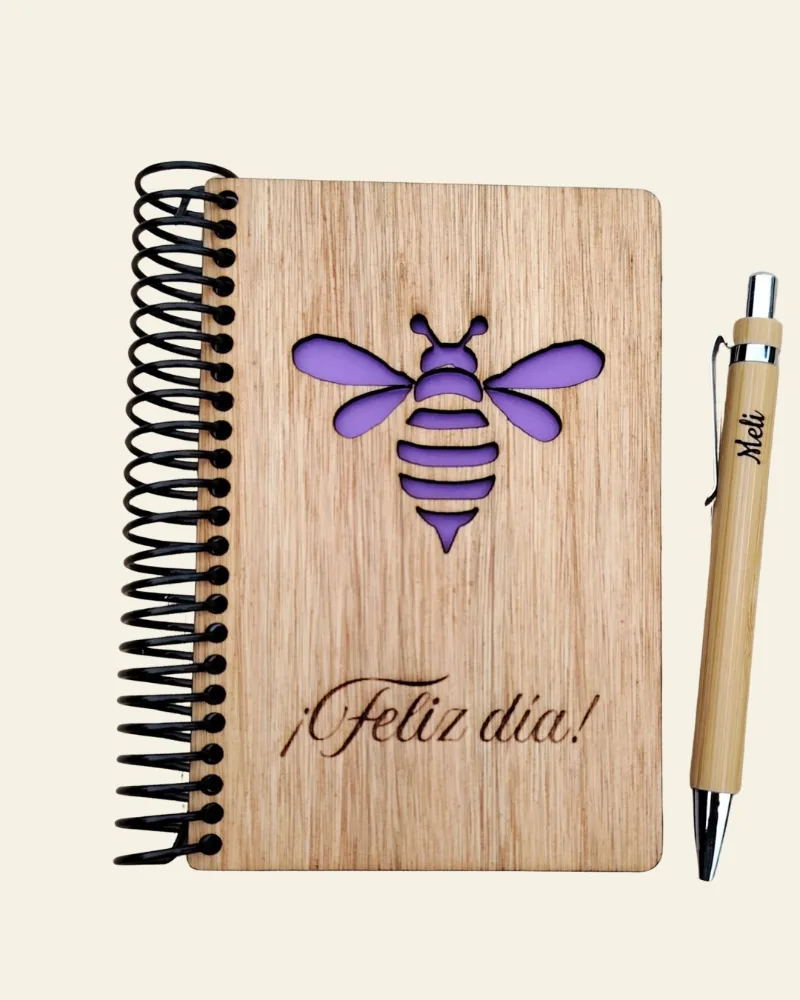 Libreta de madera con una abeja calada y papel violeta detrás, junto a una lapicera de bambú grabada con un nombre.