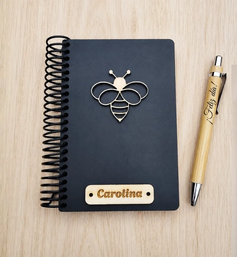 Libreta de tapa negra con una abeja de madera aplicada y una placa grabada con el nombre Carolina.