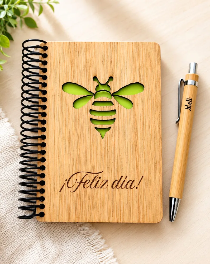 Libreta de madera con una abeja calada y papel verde detrás, grabada con la frase ¡Feliz día!