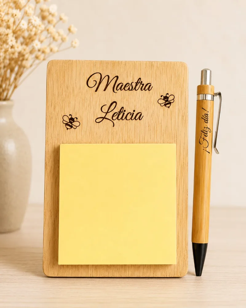 Set de escritorio de madera vertical con un taco de notas adhesivas amarillas, grabado con un nombre y dos abejas, junto a una lapicera de bambú grabada con «¡Feliz día!».