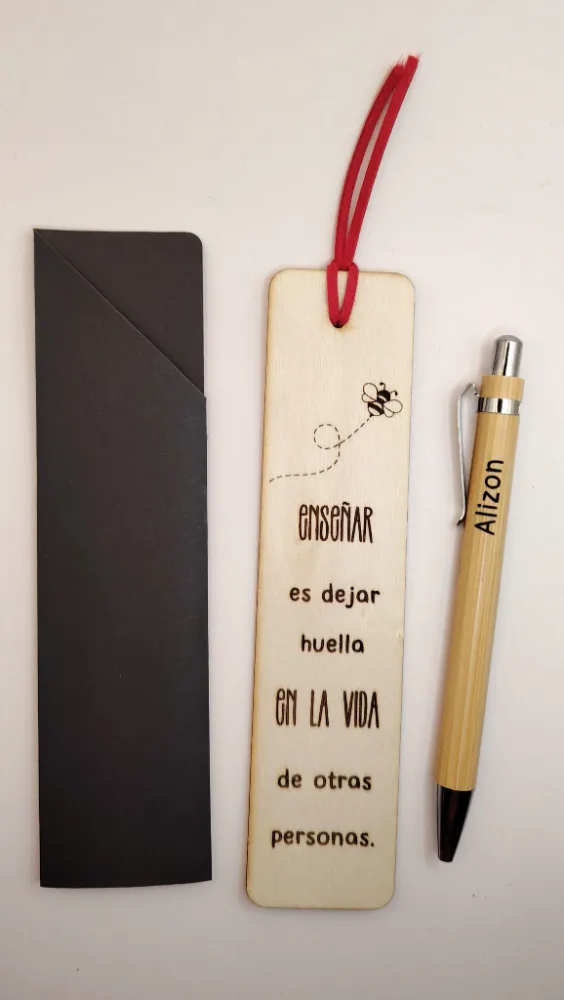 Marcalibros de madera con tiento rojo, grabado con una abeja y la frase «Enseñar es dejar huella en la vida de otras personas», entre un sobre negro y una lapicera de bambú grabada.